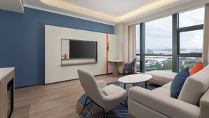 A modern hotel room with a large window, a flat-screen TV, a sofa, a small table, and a chair.