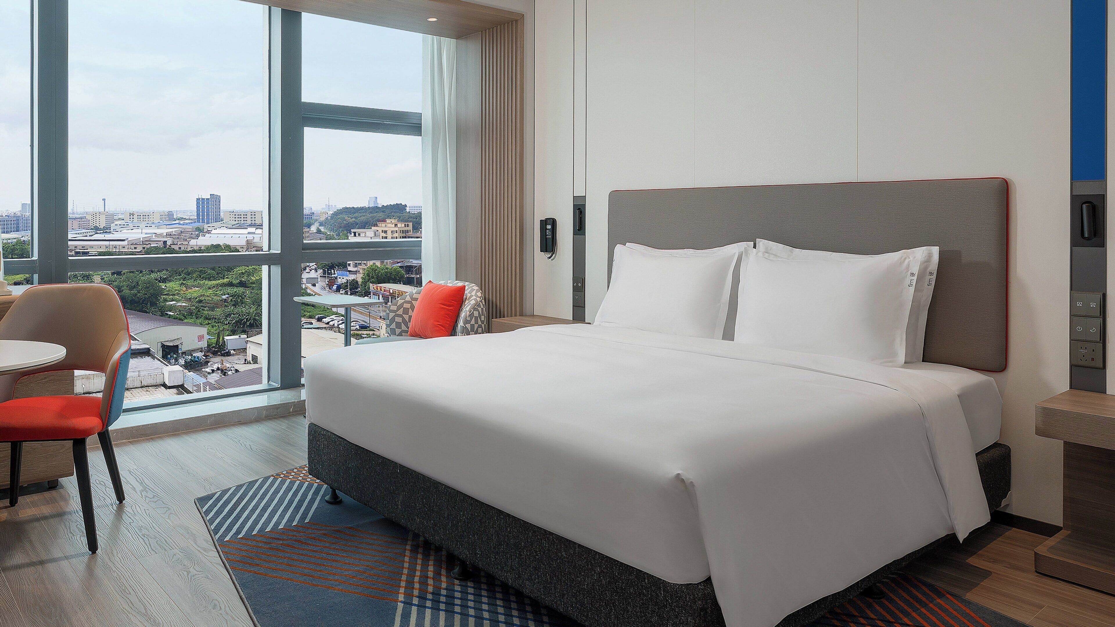 A hotel room with a large bed, a chair, a small table, and a view of the city through a large window.