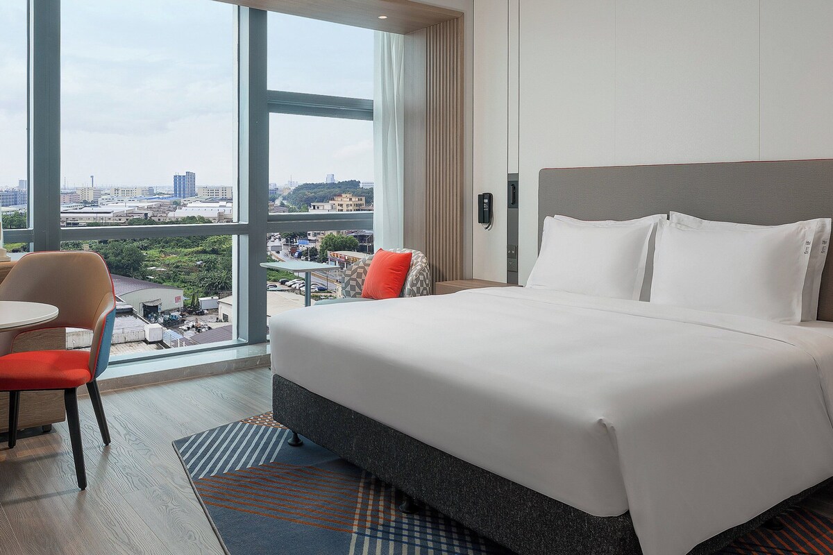 A hotel room with a large bed, a chair, a small table, and a view of the city through a large window.