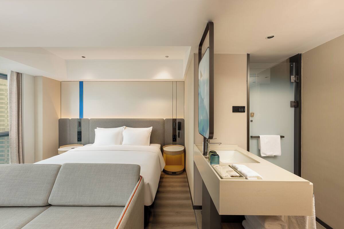 Une chambre d’hôtel moderne avec un grand lit, une table de chevet, un miroir rond, un téléviseur intégré et une salle de bain équipée d’un lavabo et d’un porte-serviettes.