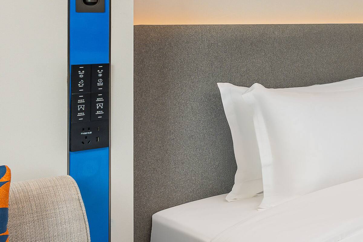 Une chambre d’hôtel avec un lit, une tête de lit grise à texture et un panneau électrique bleu orné de divers symboles et muni d’une prise de courant.