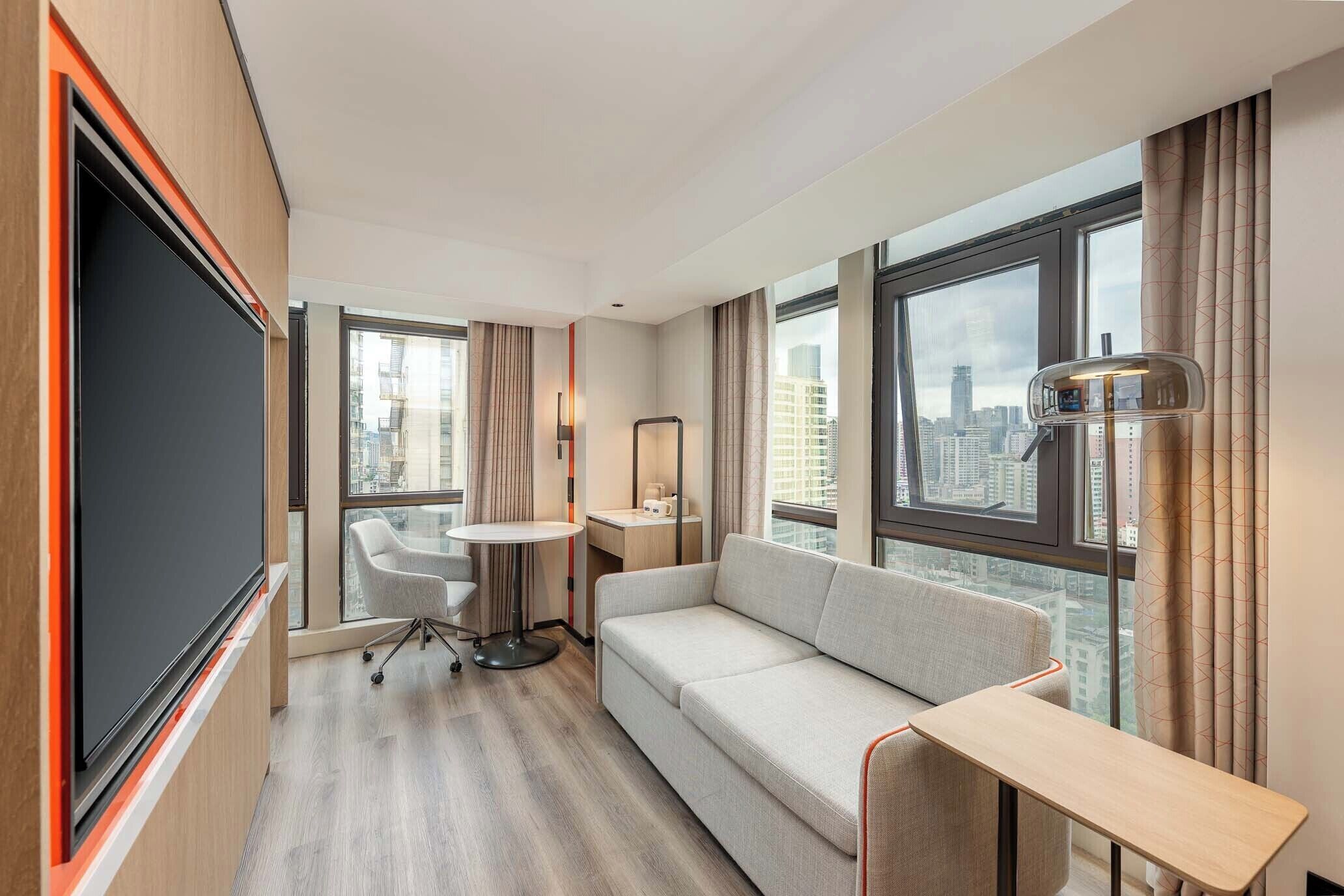 A modern hotel room with a large window, a sofa, a small table, a chair, and a flat-screen TV.