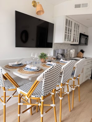 A dining area with a table set for four, surrounded by wicker chairs, a large flat-screen TV above, and a kitchen in the background.