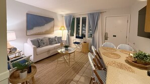 A living room with a sofa, a coffee table, a dining table with chairs, a window with curtains, and a painting on the wall. - Avalon Beach Condo (Avalon)
