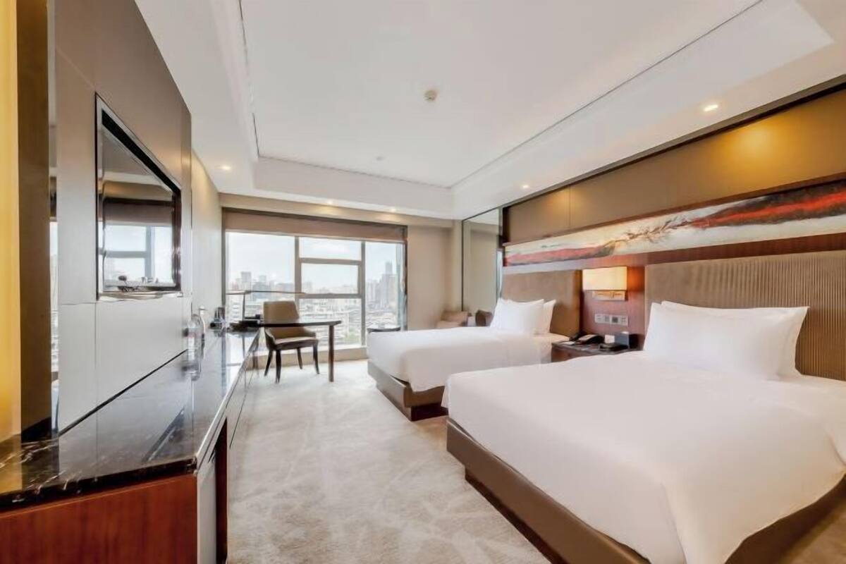 Ein Hotelzimmer mit zwei Betten, einem Schreibtisch, einem Stuhl, einem großen Fenster und Blick auf die Stadt.