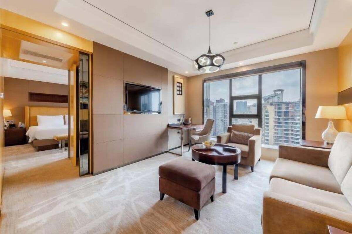 Ein modernes Hotelzimmer mit großem Fenster, einem Flachbildfernseher, einem Schreibtisch und einer gemütlichen Sitzecke mit Sofa und Sesseln.