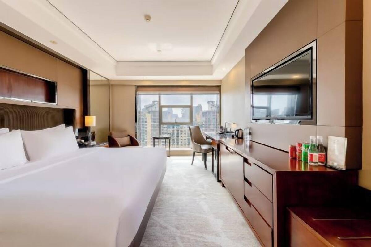 Ein Hotelzimmer mit einem großen Bett, einem Schreibtisch mit Stuhl, einem Fernseher und Blick auf die Stadt.