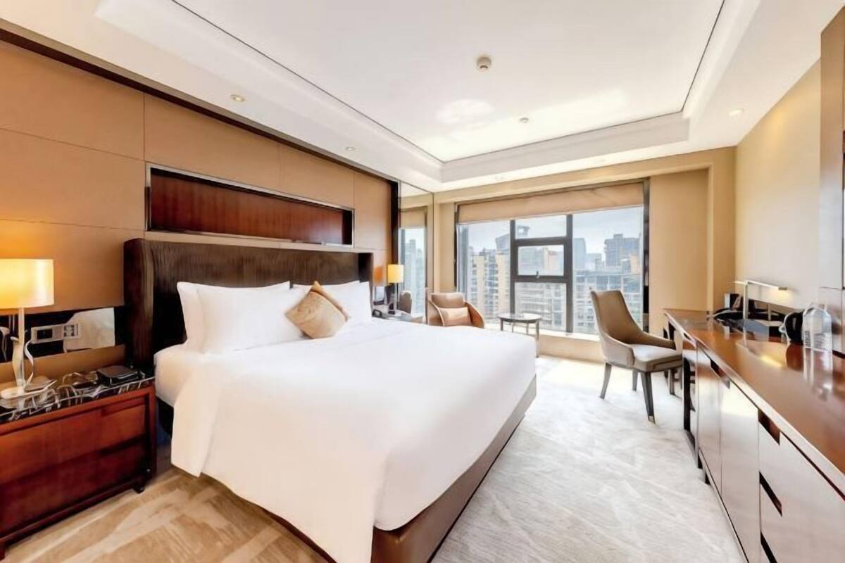 Ein Hotelzimmer mit einem großen Bett, einem Schreibtisch, einem Sessel und Blick auf die Stadt.