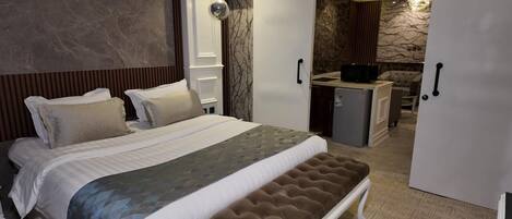 A bedroom with a large bed, a bench, a wardrobe, and a kitchenette area.