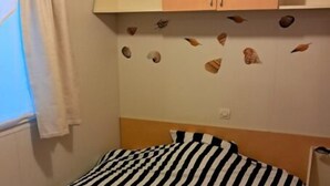 A single bed with a striped bedspread, a wooden headboard, a window with a curtain, and a wall with decorative shells. - Mobile home in the heart of nature (Minzac)