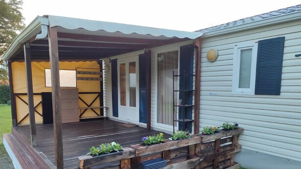 A mobile home with a covered porch, a small deck, and a window with shutters. - Mobile home in the heart of nature (Minzac)