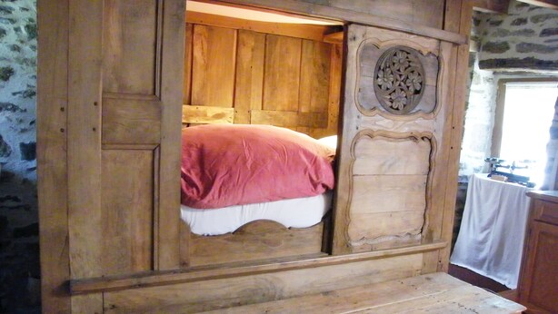 A wooden bed with a pink blanket, a wooden bench, and a decorative panel with floral carvings. - Traditional farmhouse with large garden. Porsliogan beach 500m away on foot. (Le Conquet)