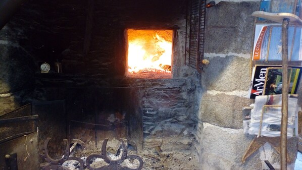 A stone fireplace with a fire burning inside, surrounded by logs and a ladder. - Traditional farmhouse with a large garden. Porsliogan Beach is a 500-meter walk away. (Le Conquet)