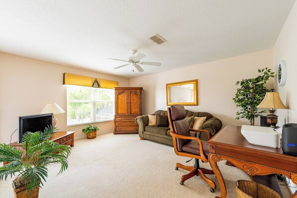 A well-lit room with a ceiling fan, a television, a desk with a computer, a sofa, and a large window with blinds.