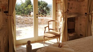 Un dormitorio con paredes de piedra, un amplio ventanal de arco, una silla de madera y una cama con cubrecama de encaje.