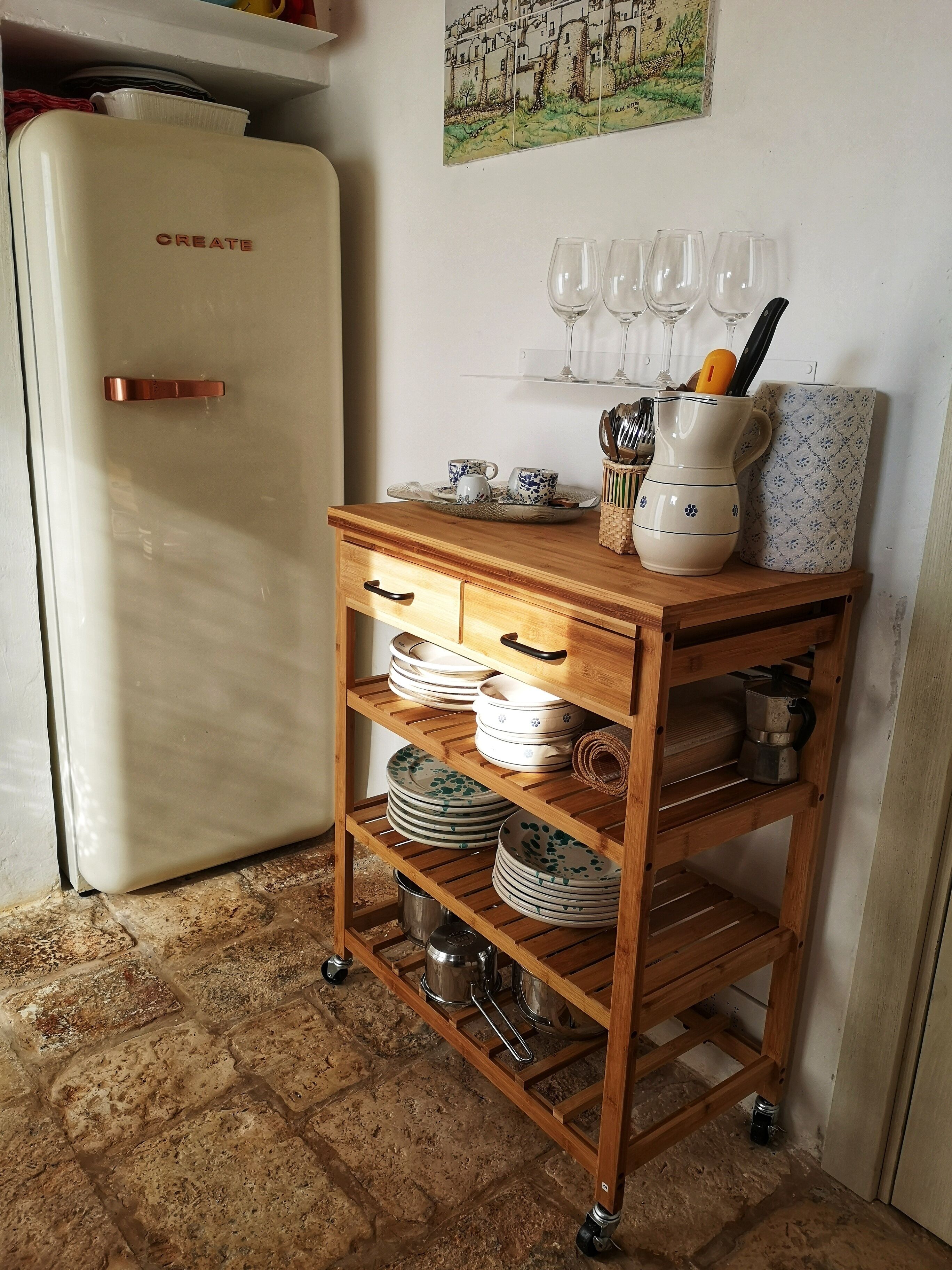 A kitchen with a white refrigerator, a wooden cart with dishes, and a wall-mounted wine rack.