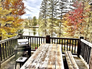 A wooden deck with a grill and picnic table overlooking a lake surrounded by autumn-colored trees.