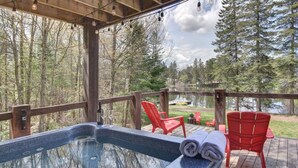 A hot tub on a wooden deck with red chairs and a view of a lake surrounded by trees.