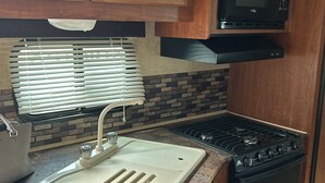 A kitchen with wooden cabinets, a stainless steel sink, a microwave, and a stove. - 1-bedroom RV in lovely Leona with a lake to fish and swim!! Exotic animals!! (Leona)