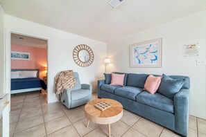 A living room with a blue sofa, a round coffee table, a grey armchair, and a bed in the background.