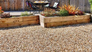 Een terras met een houten tafel en stoelen, omringd door verhoogde borders met diverse planten.