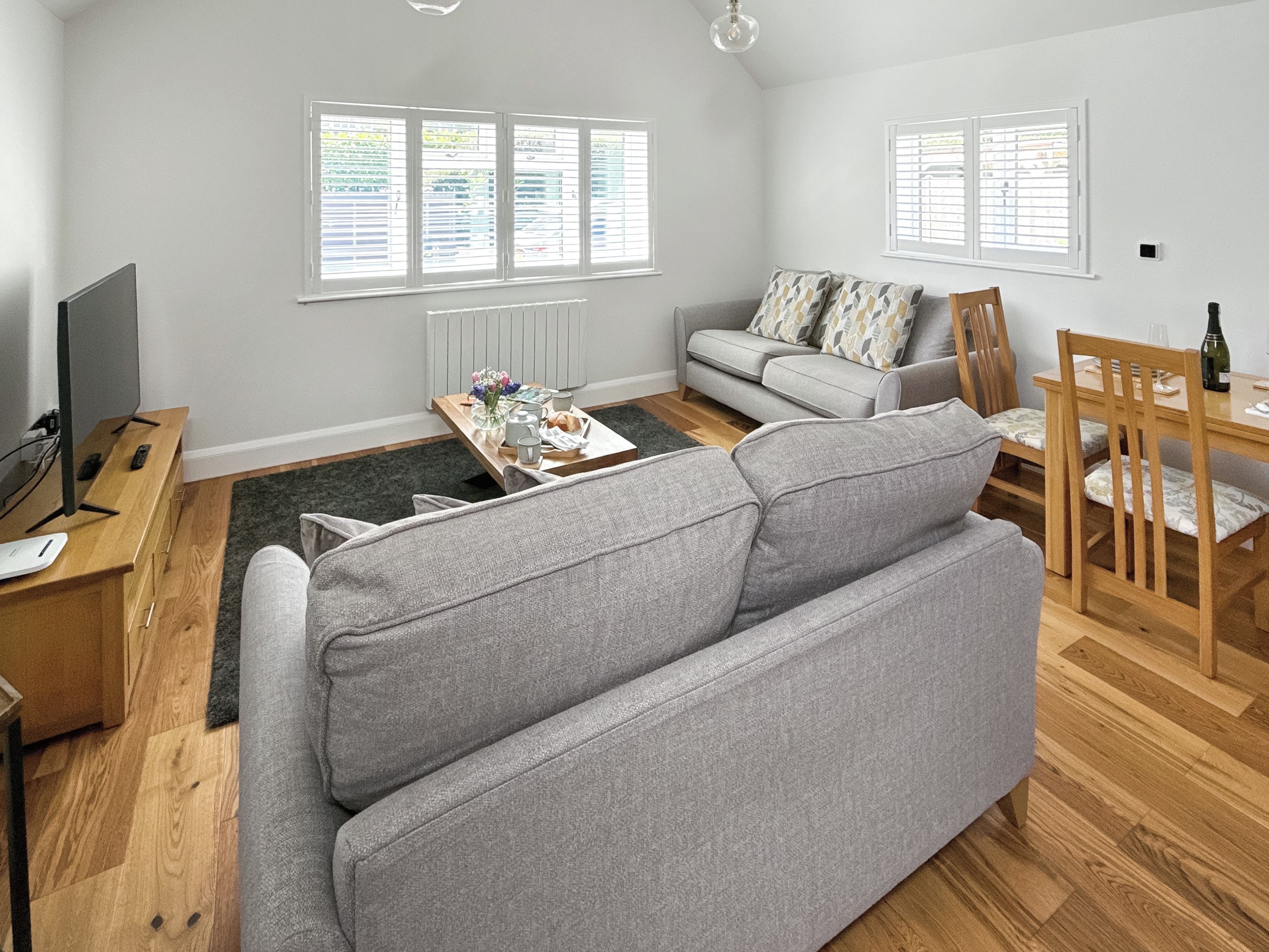 A modern living room with a grey sofa, a wooden dining table with chairs, a TV on a wooden stand, and a radiator.