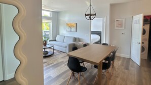 A modern living room with a dining area, hardwood flooring, and a large window providing natural light.