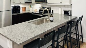 A modern kitchen with white cabinets, a granite countertop, a stainless steel refrigerator, a microwave, a toaster, and a bar stool area.