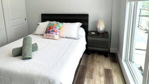 A neatly made bed with a white comforter and pillows, a bedside lamp, and a window with a view of greenery.