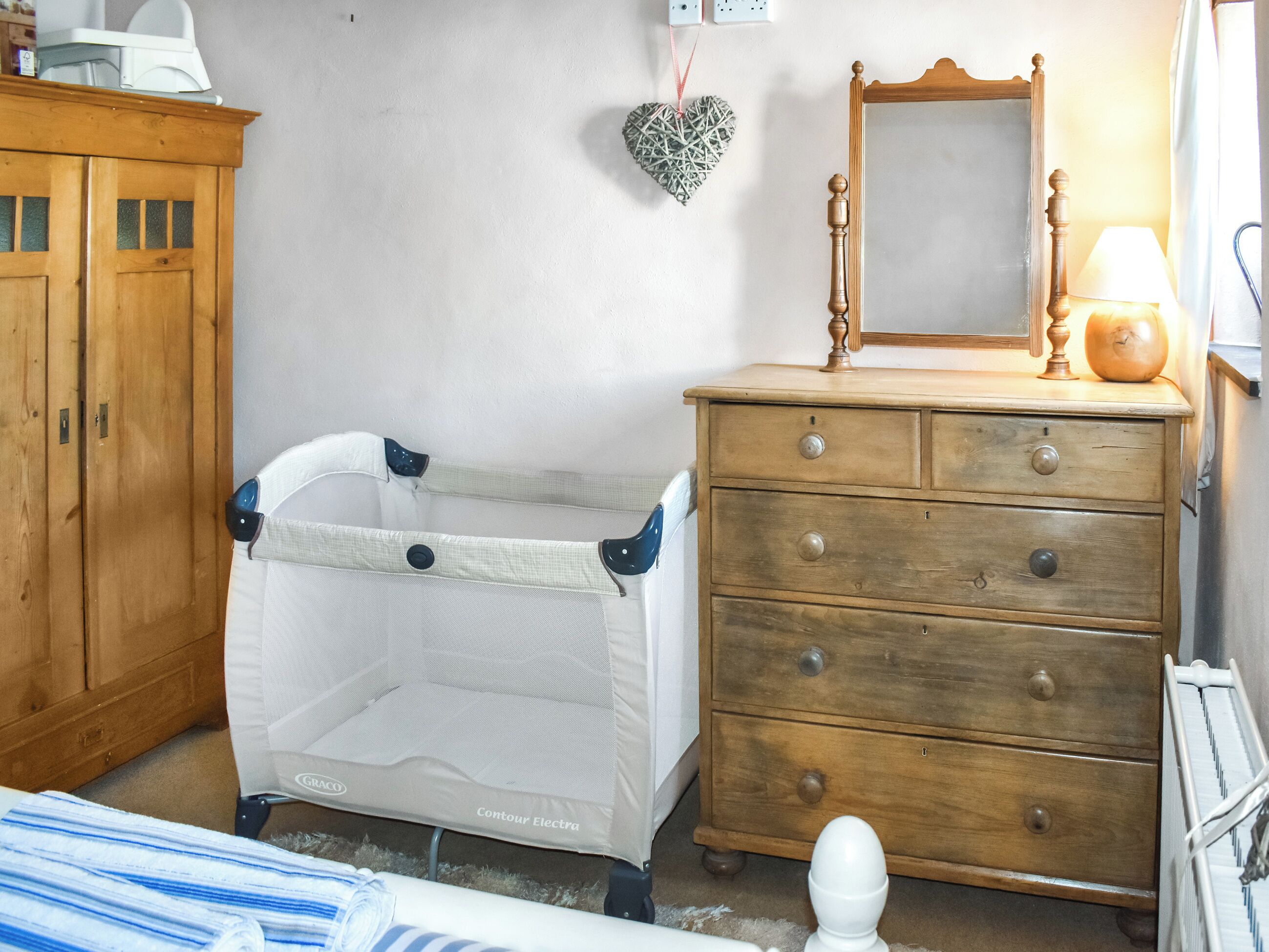 A room with a wooden cabinet, a baby crib, a dresser with drawers, a mirror, and a lamp.
