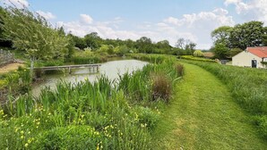 A serene riverside pathway with a wooden bridge, lush greenery, and a small building with a red roof.