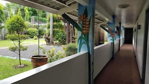 A covered corridor with a tiled floor, a view of a garden with trees and a pathway, and a mural on the wall.