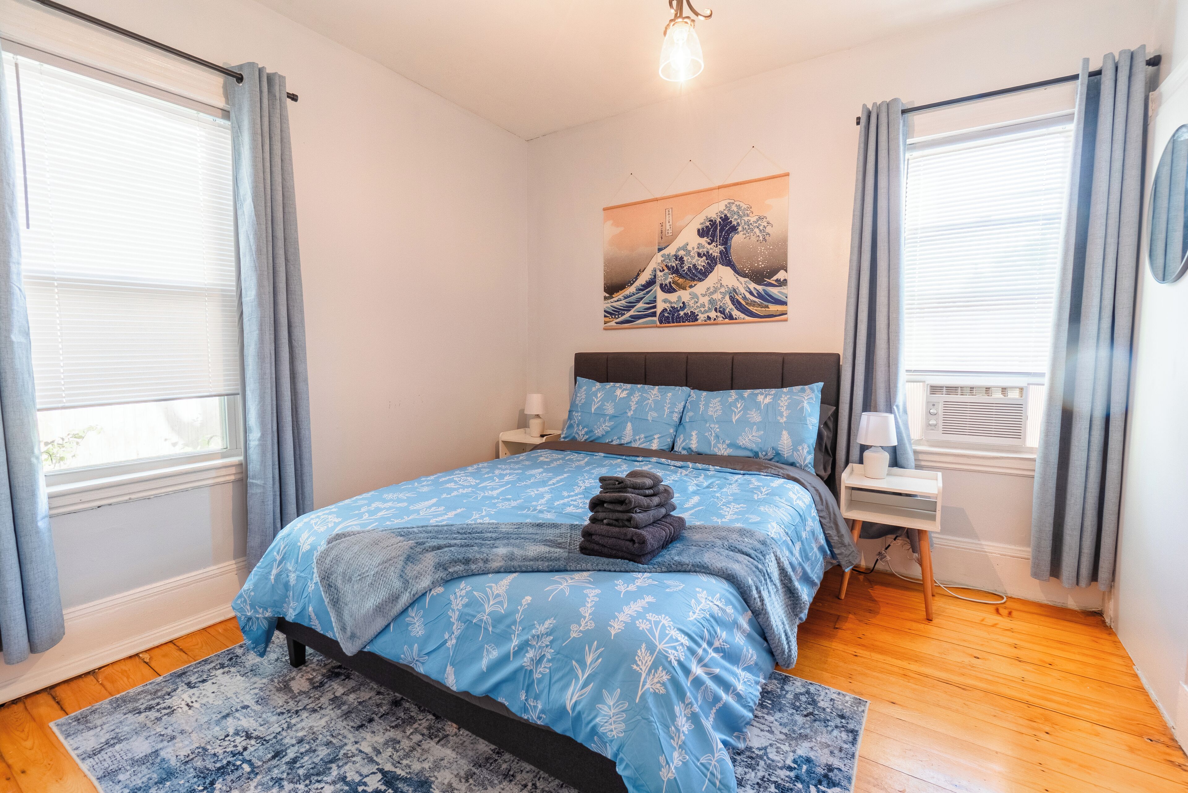 A bedroom with a bed, a nightstand, a lamp, a painting, and a window with curtains.
