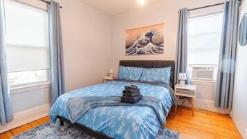 A bedroom with a bed, a nightstand, a lamp, a painting, and a window with curtains.