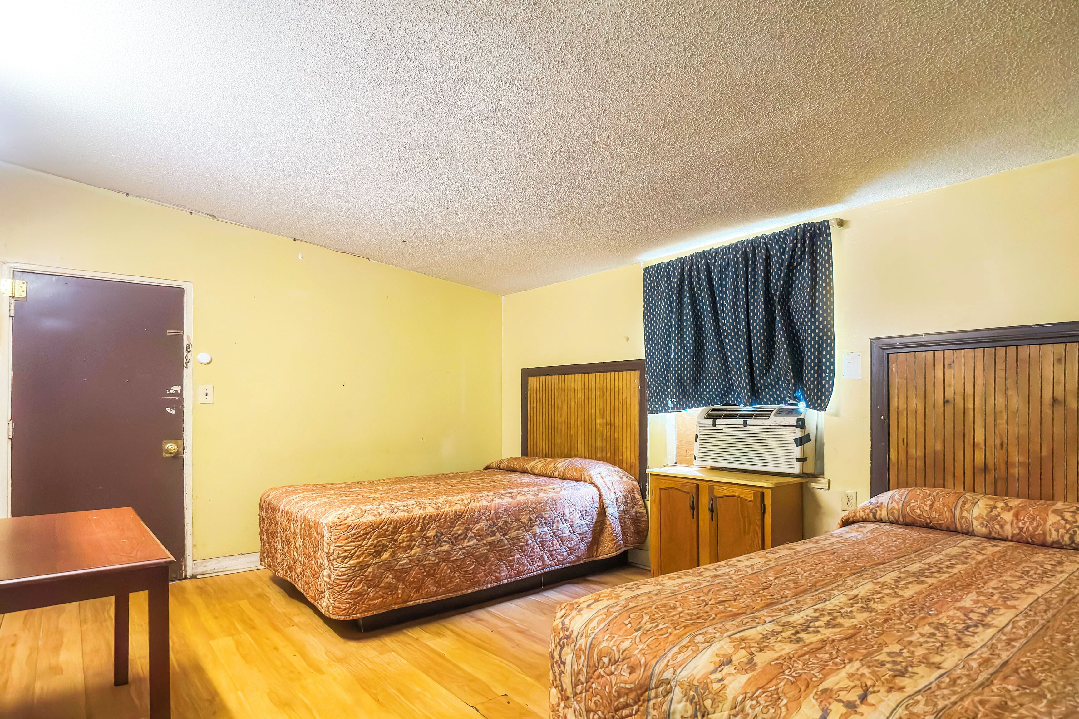 A room with two beds, a wooden table, a door, and a window with curtains.