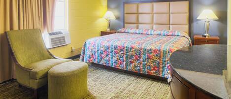 A hotel room with a large bed, a chair, an ottoman, a nightstand with a lamp, and an air conditioning unit.