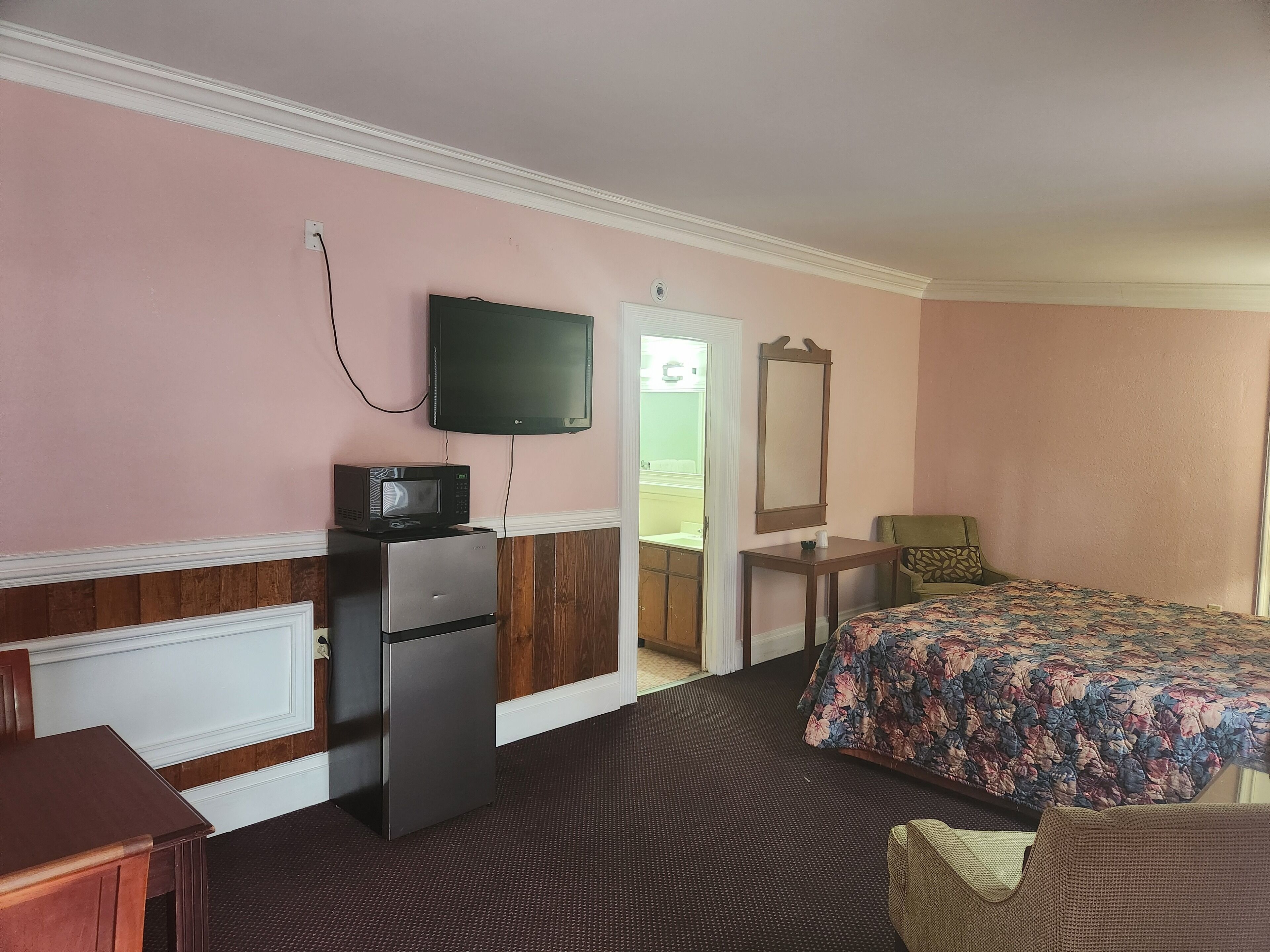 A hotel room with a bed, a television, a refrigerator, a wooden cabinet, a mirror, and a door leading to a bathroom.