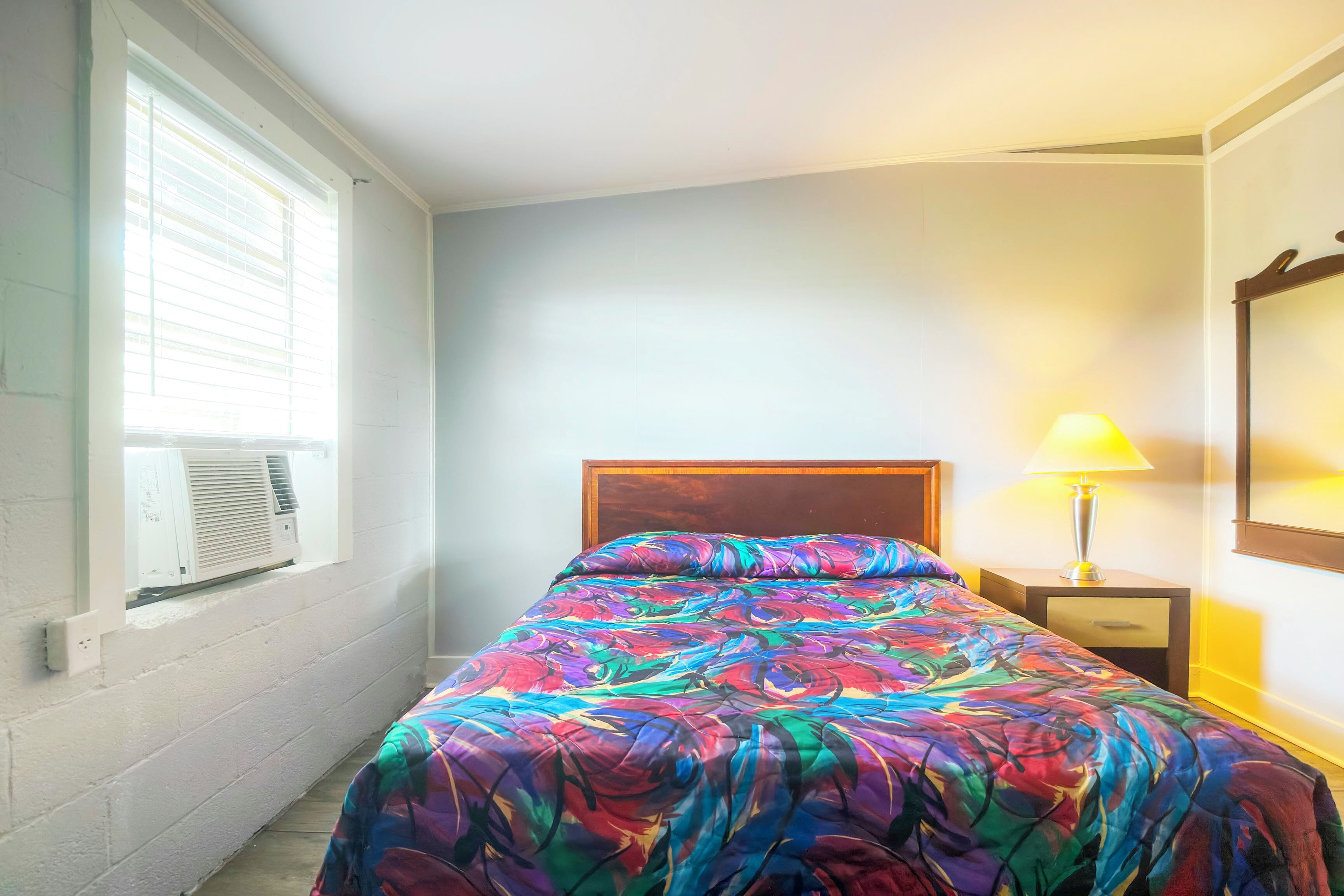 Un dormitorio con cama, lámpara de noche, ventana con persianas y aire acondicionado.