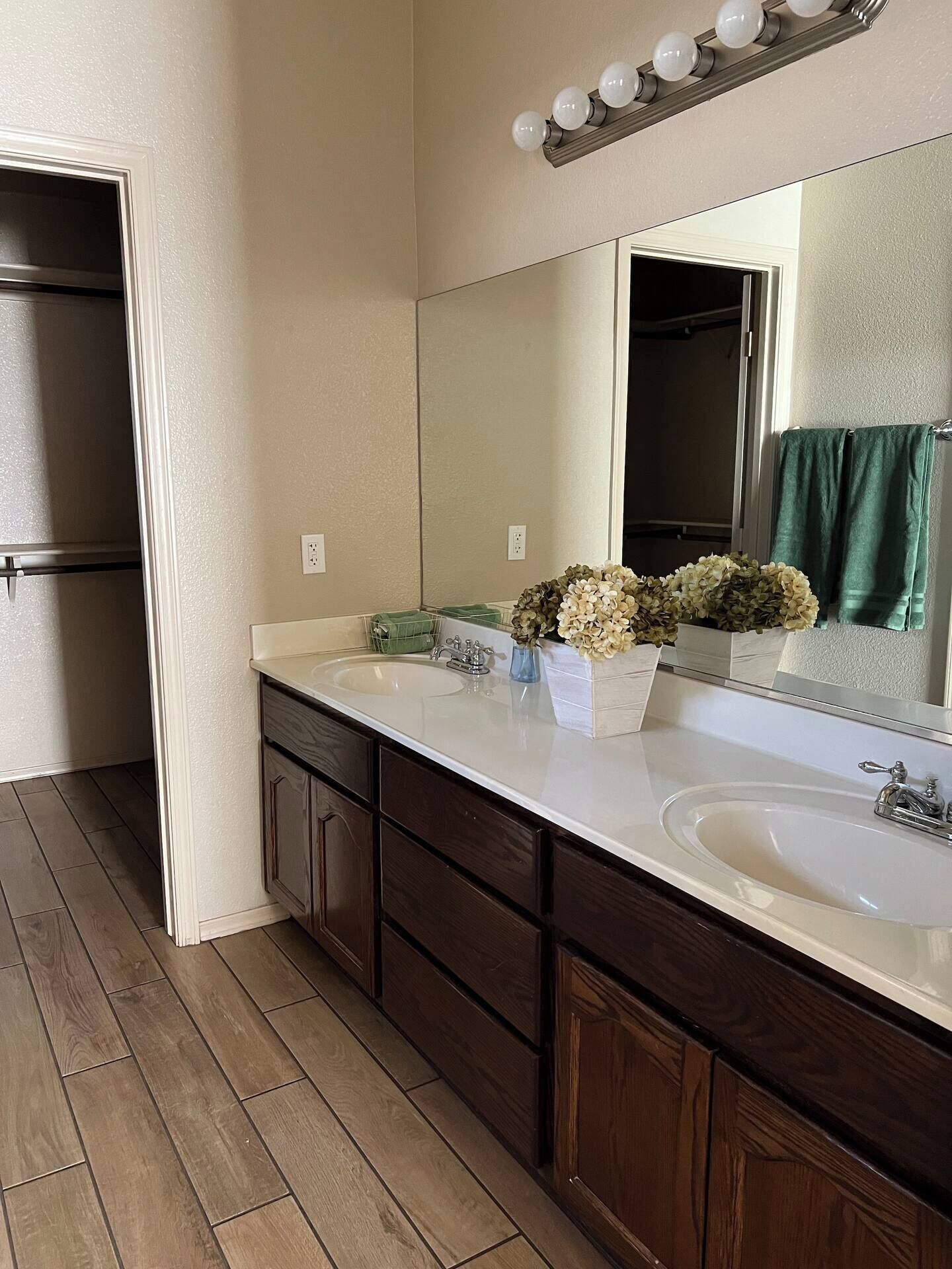 Une salle de bain avec un double lavabo, des meubles en bois, un miroir avec éclairage intégré et une serviette verte suspendue.