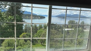 A large window with a grid pattern, offering a view of a lake, mountains, and surrounding greenery. - New! Charming Blue Haven Cottage with Amazing Lake Pend Oreille Views! (Hope)