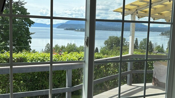 A view from a covered deck with a railing, overlooking a body of water and greenery. - New! Charming Blue Haven Cottage with Amazing Lake Pend Oreille Views! (Hope)