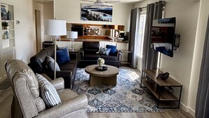 A modern living room with a grey leather sofa, a black leather armchair, and a round coffee table. There is a flat-screen TV mounted on the wall, a ceiling fan, and a kitchen area visible through an open door. - New! Charming Blue Haven Cottage with Amazing Lake Pend Oreille Views! (Hope)