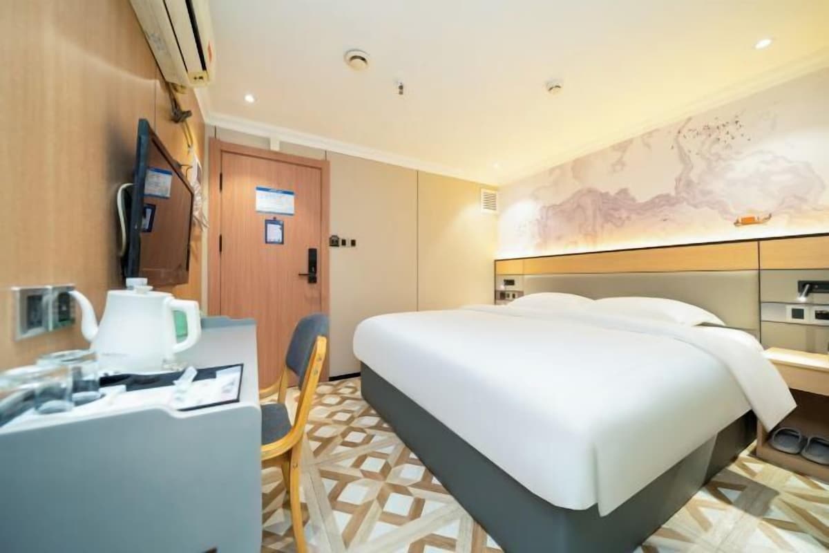 A hotel room with a bed, a desk with a kettle, a chair, a TV, and a wall with a painting.