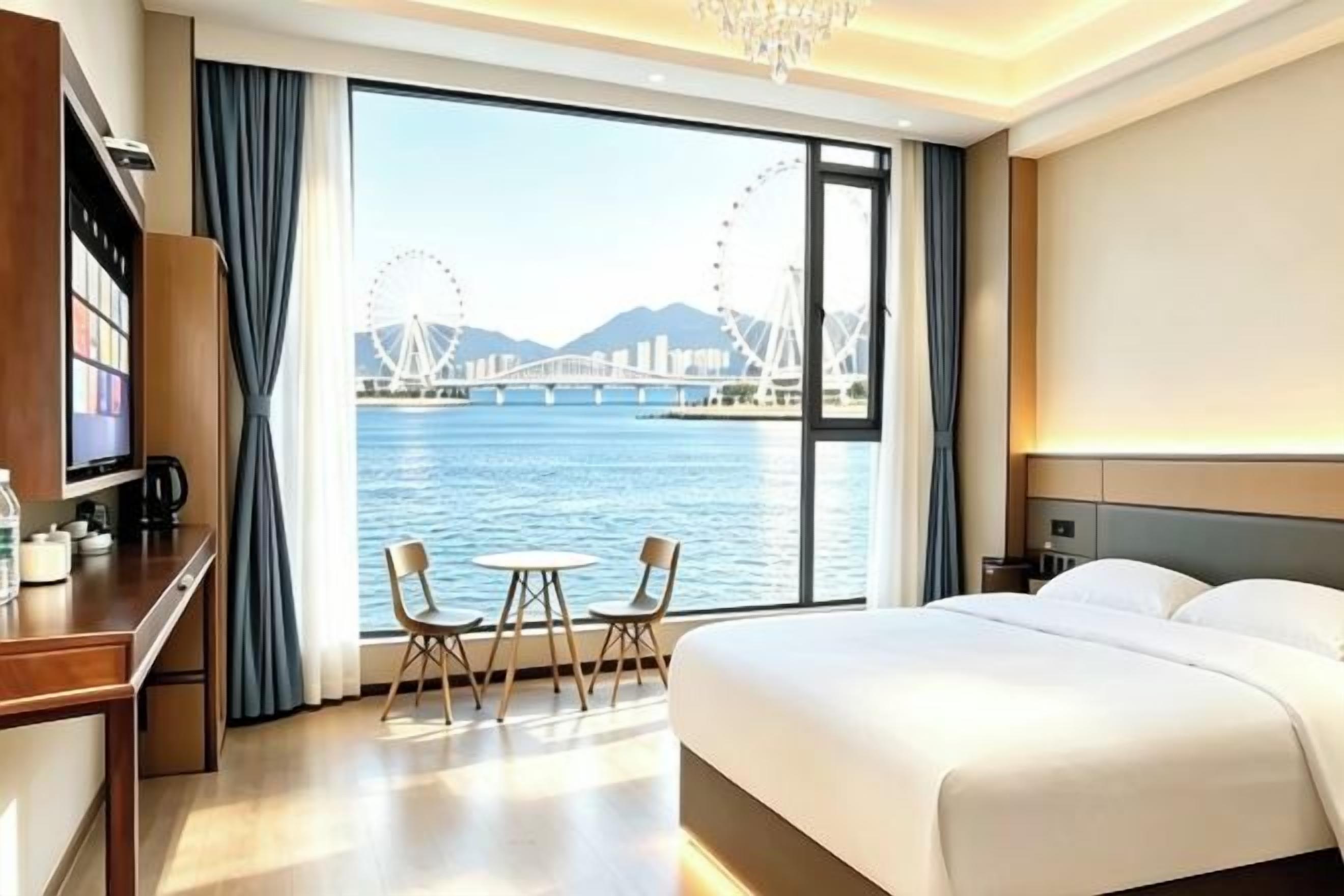 A hotel room with a large window, a bed, a desk with a chair, and a small table.