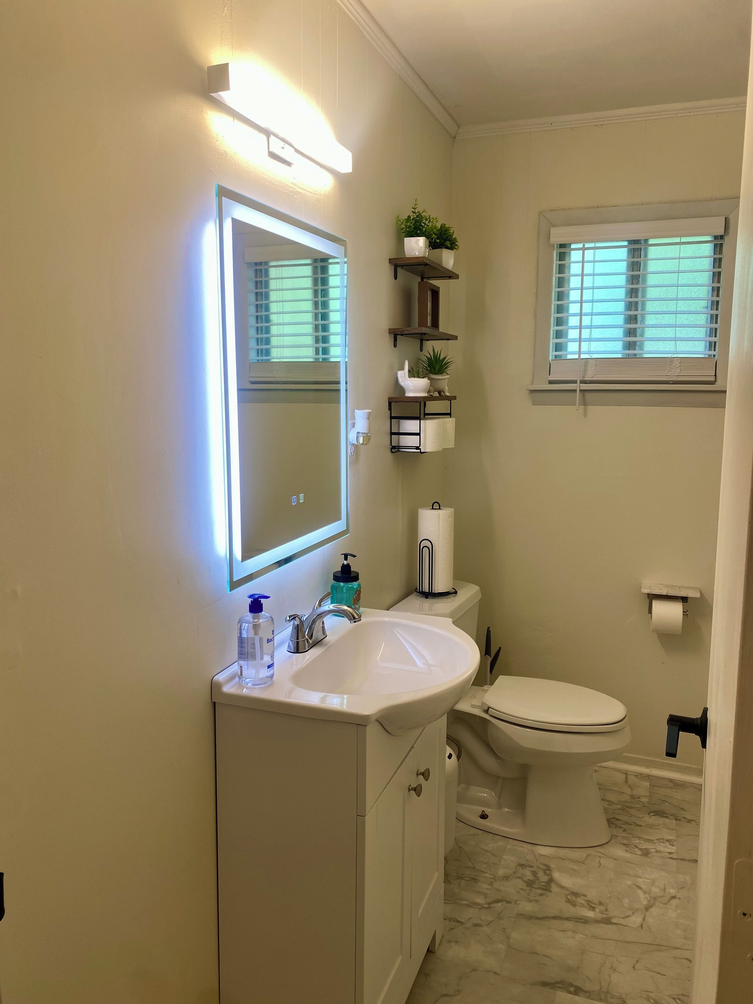 Un baño con lavamanos blanco, espejo, inodoro y ventana con persianas.