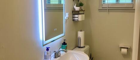 Un baño con lavamanos blanco, espejo, inodoro y ventana con persianas.