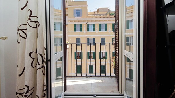 A view through a balcony door with a patterned curtain, leading to a building with multiple windows and balconies.