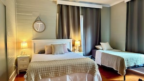 A hotel room with a large bed, a smaller bed, two bedside tables with lamps, a wall-mounted picture frame, and a window with curtains.