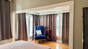 Un dormitorio con cama, un sillón azul con almohada blanca, una mesita con lámpara y ventanas grandes con cortinas.
