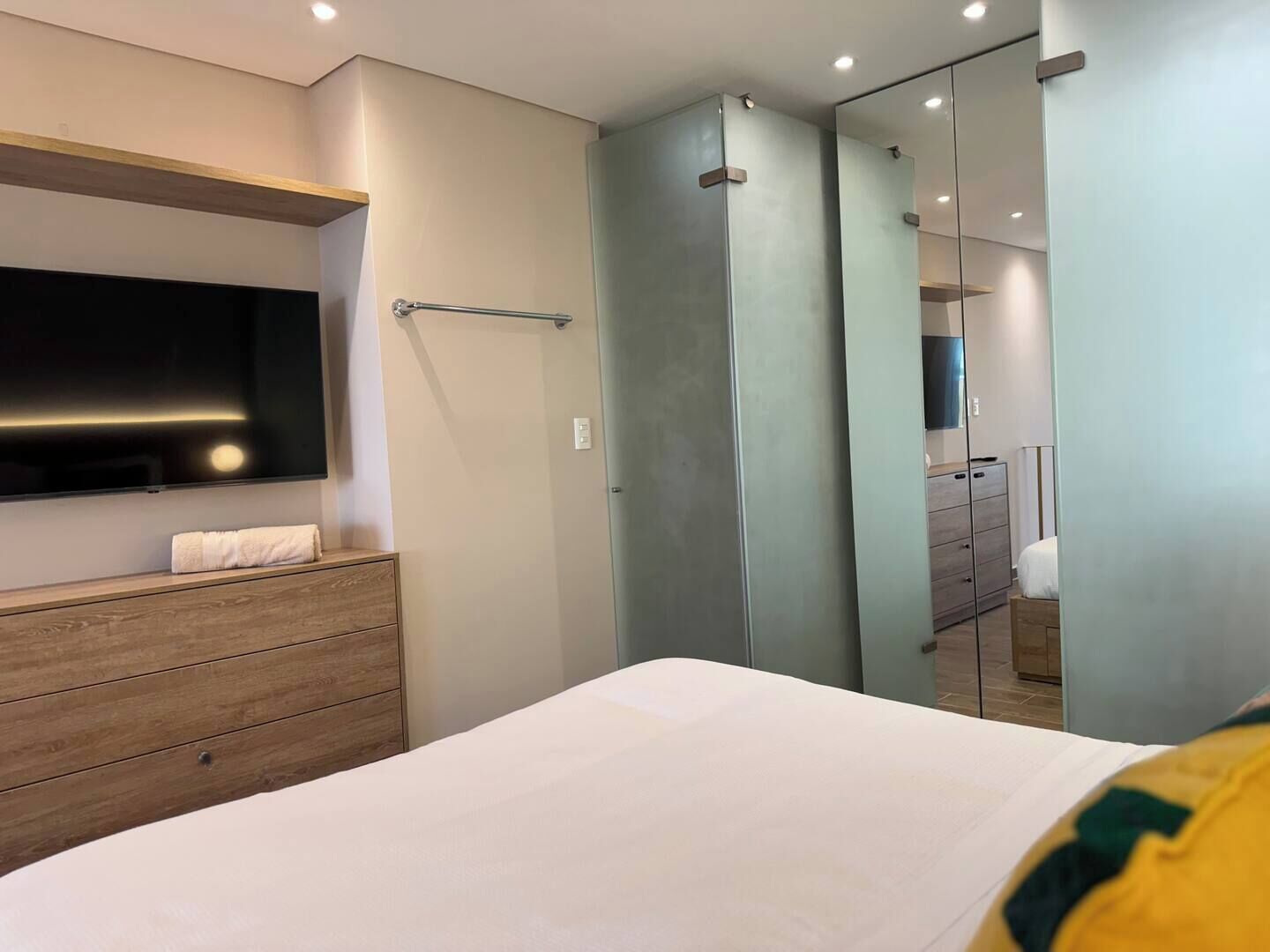 A modern bedroom with a large bed, a wooden dresser, a flat-screen TV mounted on the wall, and a mirror cabinet.
